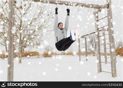 fitness, sport, exercising, training and people concept - young man doing leg pull ups on horizontal bar and flexing abdominal muscles outdoors in winter. young man exercising on horizontal bar in winter