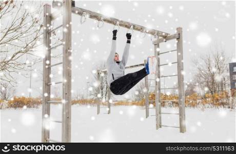 fitness, sport, exercising, training and people concept - young man doing leg pull ups on horizontal bar and flexing abdominal muscles outdoors in winter. young man exercising on horizontal bar in winter