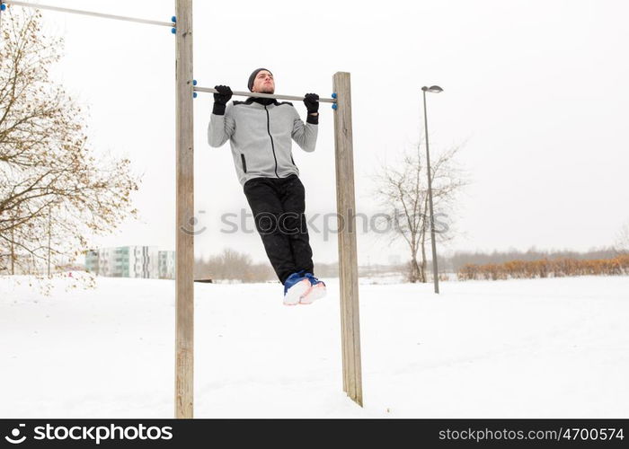 fitness, sport, exercising, training and people concept - young man doing pull ups on horizontal bar outdoors in winter