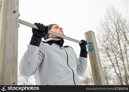 fitness, sport, exercising, training and people concept - young man doing pull ups on horizontal bar outdoors in winter