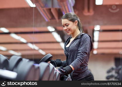 fitness, sport, exercising, weightlifting and people concept - young smiling woman choosing dumbbells in gym