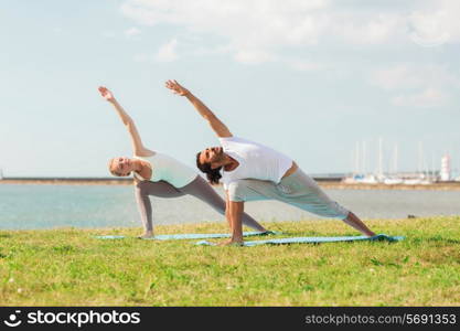 fitness, sport, friendship and lifestyle concept - smiling couple making yoga exercises on mats outdoors