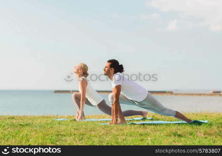 fitness, sport, friendship and lifestyle concept - smiling couple making yoga exercises on mats outdoors