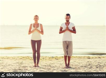 fitness, sport, friendship and lifestyle concept - smiling couple making yoga exercises on sand outdoors