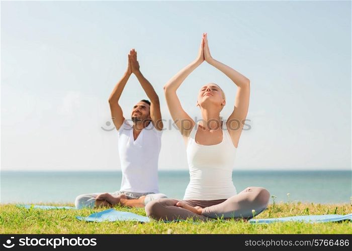 fitness, sport, friendship and lifestyle concept - smiling couple making yoga exercises sitting on mats outdoors