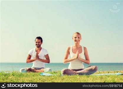 fitness, sport, friendship and lifestyle concept - smiling couple making yoga exercises sitting on mats outdoors