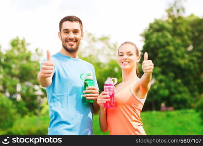fitness, sport, friendship and lifestyle concept - smiling couple with bottles of water showing thumbs up outdoors