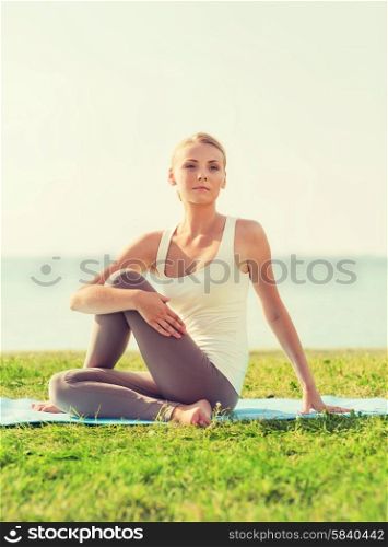 fitness, sport, friendship and lifestyle concept - smiling woman making yoga exercises sitting on mat outdoors