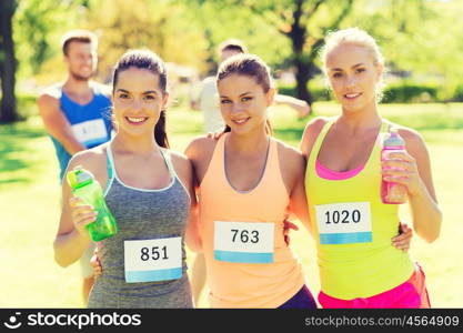 fitness, sport, friendship, marathon and healthy lifestyle concept - happy young sporty women with racing badge numbers and water bottles outdoors