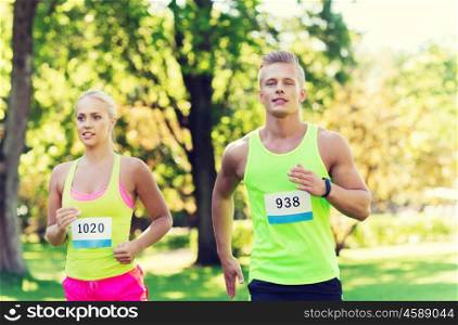fitness, sport, friendship, race and healthy lifestyle concept - happy teenage friends or sportsmen couple running marathon with badge numbers outdoors