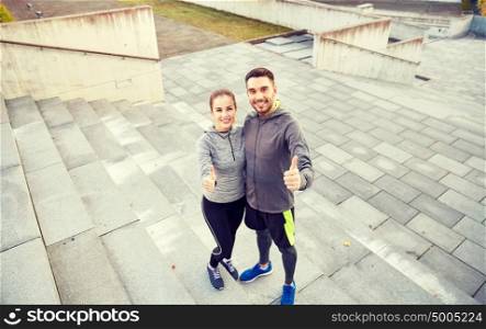 fitness, sport, people and gesture concept - smiling couple outdoors showing thumbs up on city street stairs. smiling couple showing thumbs up on city street