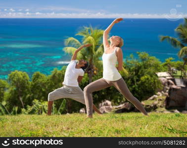 fitness, sport, people and healthy lifestyle concept - couple doing yoga in warrior pose over natural background and sea. couple doing yoga over natural background and sea