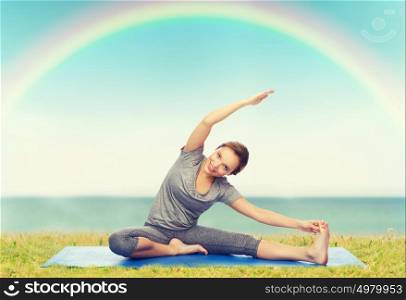 fitness, sport, people and healthy lifestyle concept - happy woman making yoga and stretching on mat over blue sky, rainbow and sea background. happy woman making yoga and stretching on mat
