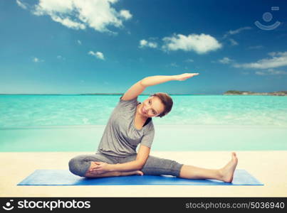 fitness, sport, people and healthy lifestyle concept - happy woman making yoga and stretching on mat over beach background. happy woman making yoga and stretching on mat