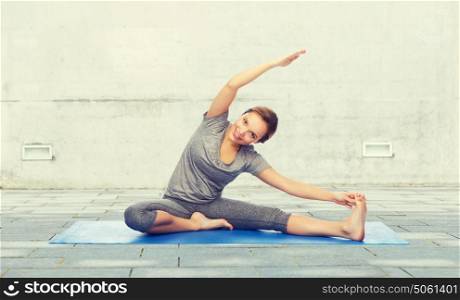 fitness, sport, people and healthy lifestyle concept - happy woman making yoga and stretching on mat over urban street background. happy woman making yoga and stretching on mat