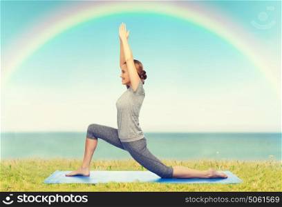 fitness, sport, people and healthy lifestyle concept - happy woman making yoga in low lunge pose on mat over blue sky, rainbow and sea background. happy woman making yoga in low lunge on mat