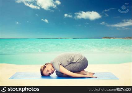 fitness, sport, people and healthy lifestyle concept - happy woman making yoga in child pose on mat over sea and sky background. happy woman making yoga in child pose on beach