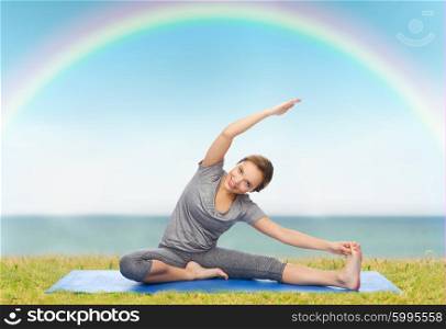 fitness, sport, people and healthy lifestyle concept - happy woman making yoga and stretching on mat over blue sky, rainbow and sea background