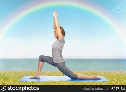 fitness, sport, people and healthy lifestyle concept - happy woman making yoga in low lunge pose on mat over blue sky, rainbow and sea background