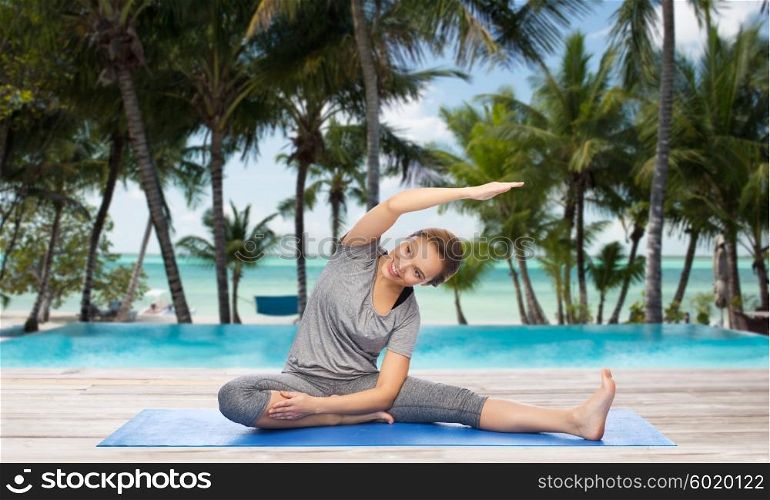 fitness, sport, people and healthy lifestyle concept - happy woman making yoga and stretching on mat over hotel resort pool on tropical beach background