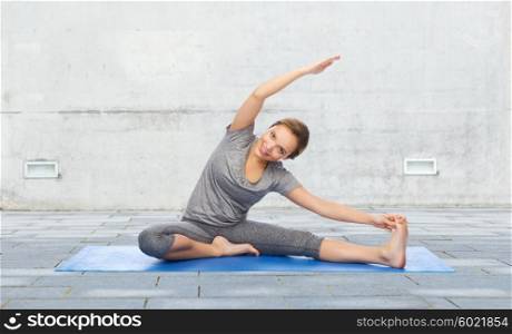 fitness, sport, people and healthy lifestyle concept - happy woman making yoga and stretching on mat over urban street background