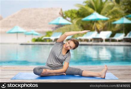 fitness, sport, people and healthy lifestyle concept - happy woman making yoga and stretching on mat over beach and swimming pool background