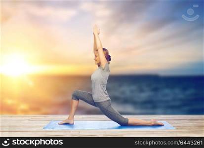 fitness, sport, people and healthy lifestyle concept - happy woman making yoga in low lunge pose on pier over sea background. happy woman making yoga in low lunge on mat