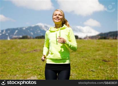 fitness, sport, people and healthy lifestyle concept - happy young smiling woman jogging or running over mountains, green field and blue sky background