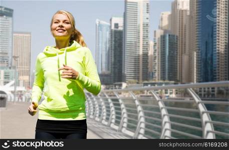 fitness, sport, people and healthy lifestyle concept - happy young woman jogging over dubai city street or waterfront background