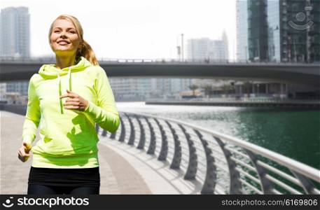 fitness, sport, people and healthy lifestyle concept - happy young woman jogging over dubai city street or waterfront and bridge background