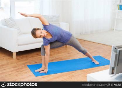 fitness, sport, people and healthy lifestyle concept - woman making yoga bikram triangle pose on mat and watching tv lesson