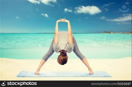 fitness, sport, people and healthy lifestyle concept - woman making yoga in wide-legged forward bend pose on mat over sea and sky background. woman making yoga forward bend on beach