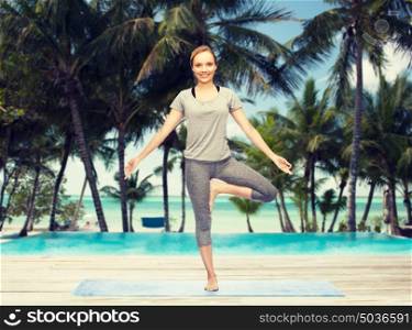 fitness, sport, people and healthy lifestyle concept - woman making yoga in tree pose on mat over hotel resort pool on tropical beach background. woman making yoga in tree pose on mat
