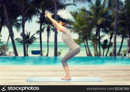 fitness, sport, people and healthy lifestyle concept - woman making yoga in chair pose on mat over hotel resort pool on tropical beach background. woman making yoga in chair pose on mat