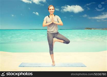 fitness, sport, people and healthy lifestyle concept - woman making yoga in tree pose on mat over sea and sky background. woman making yoga in tree pose on beach