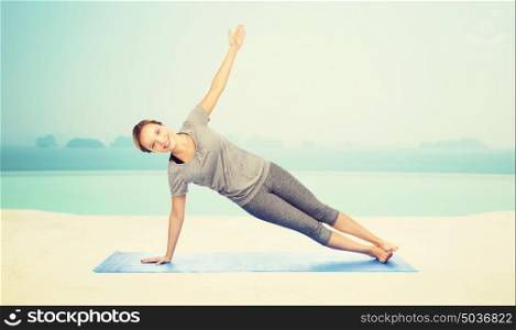 fitness, sport, people and healthy lifestyle concept - woman making yoga in side plank pose on mat over infinity edge pool at hotel resort background. woman making yoga in side plank pose on mat