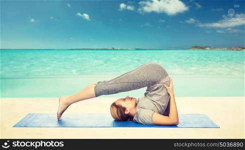 fitness, sport, people and healthy lifestyle concept - woman making yoga in plow pose on mat over beach background. woman making yoga in plow pose on mat