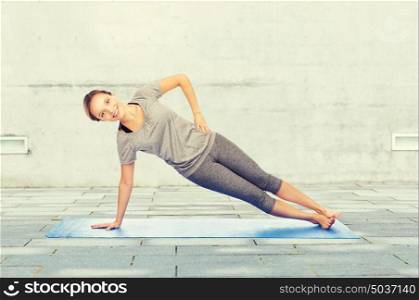 fitness, sport, people and healthy lifestyle concept - woman making yoga in side plank pose on mat over urban street background. woman making yoga in side plank pose on mat