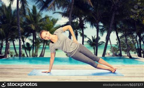 fitness, sport, people and healthy lifestyle concept - woman making yoga in side plank pose on mat over hotel resort pool on tropical beach background. woman making yoga in side plank pose on mat