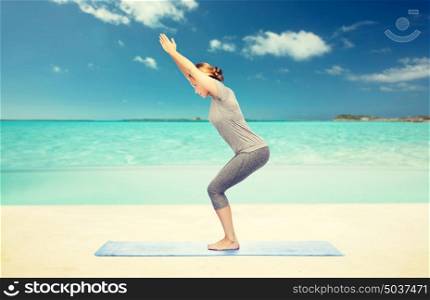 fitness, sport, people and healthy lifestyle concept - woman making yoga in chair pose on mat over beach background. woman making yoga in chair pose on mat