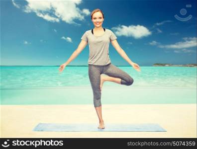fitness, sport, people and healthy lifestyle concept - woman making yoga in tree pose on mat over beach background. woman making yoga in tree pose on mat