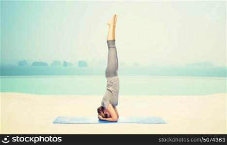 fitness, sport, people and healthy lifestyle concept - woman making yoga in headstand pose on mat over infinity edge pool at hotel resort background. woman making yoga in headstand pose on mat