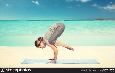 fitness, sport, people and healthy lifestyle concept - woman making yoga in crane pose on mat over sea and sky background. woman making yoga in crane pose on mat over beach
