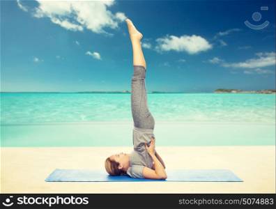 fitness, sport, people and healthy lifestyle concept - woman making yoga in shoulderstand pose on mat over beach background. woman making yoga in shoulderstand pose on mat