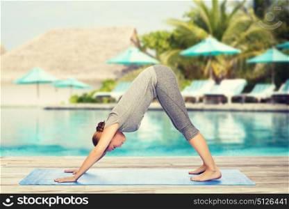 fitness, sport, people and healthy lifestyle concept - woman making yoga in downward facing dog pose on mat over beach and swimming pool background. woman making yoga dog pose on mat
