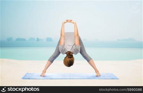 fitness, sport, people and healthy lifestyle concept - woman making yoga in wide-legged forward bend pose on mat over infinity edge pool at hotel resort background
