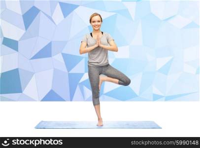 fitness, sport, people and healthy lifestyle concept - woman making yoga in tree pose on mat over blue polygonal background