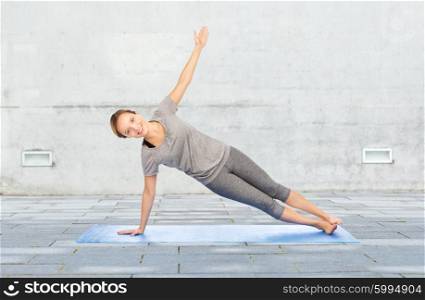 fitness, sport, people and healthy lifestyle concept - woman making yoga in side plank pose on mat over urban street background