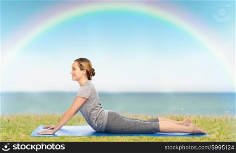 fitness, sport, people and healthy lifestyle concept - woman making yoga in dog pose on mat over blue sky, rainbow and sea background