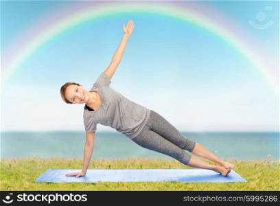 fitness, sport, people and healthy lifestyle concept - woman making yoga in side plank pose on mat over blue sky, rainbow and sea background